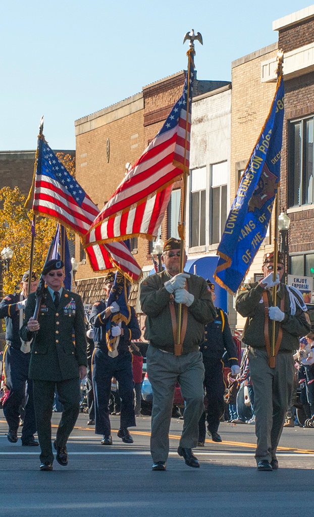 A photo taken during the Veterans Day parade in Emporia, Kansas. It shows servicemen marching down Comercial Street while they hold flags.